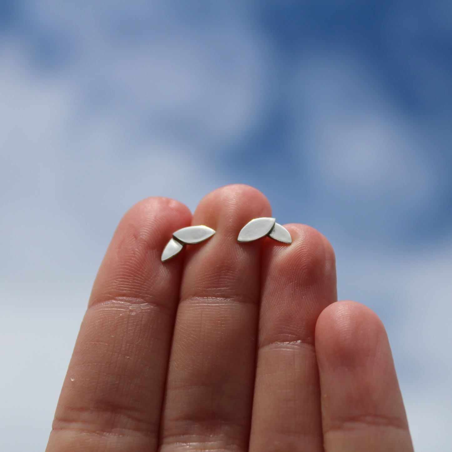 A pair of silver shiny finished two leaf stud earrings held in a hand against a sky background. Handmade from Sterling Silver by Jen Lithgo Jewellery