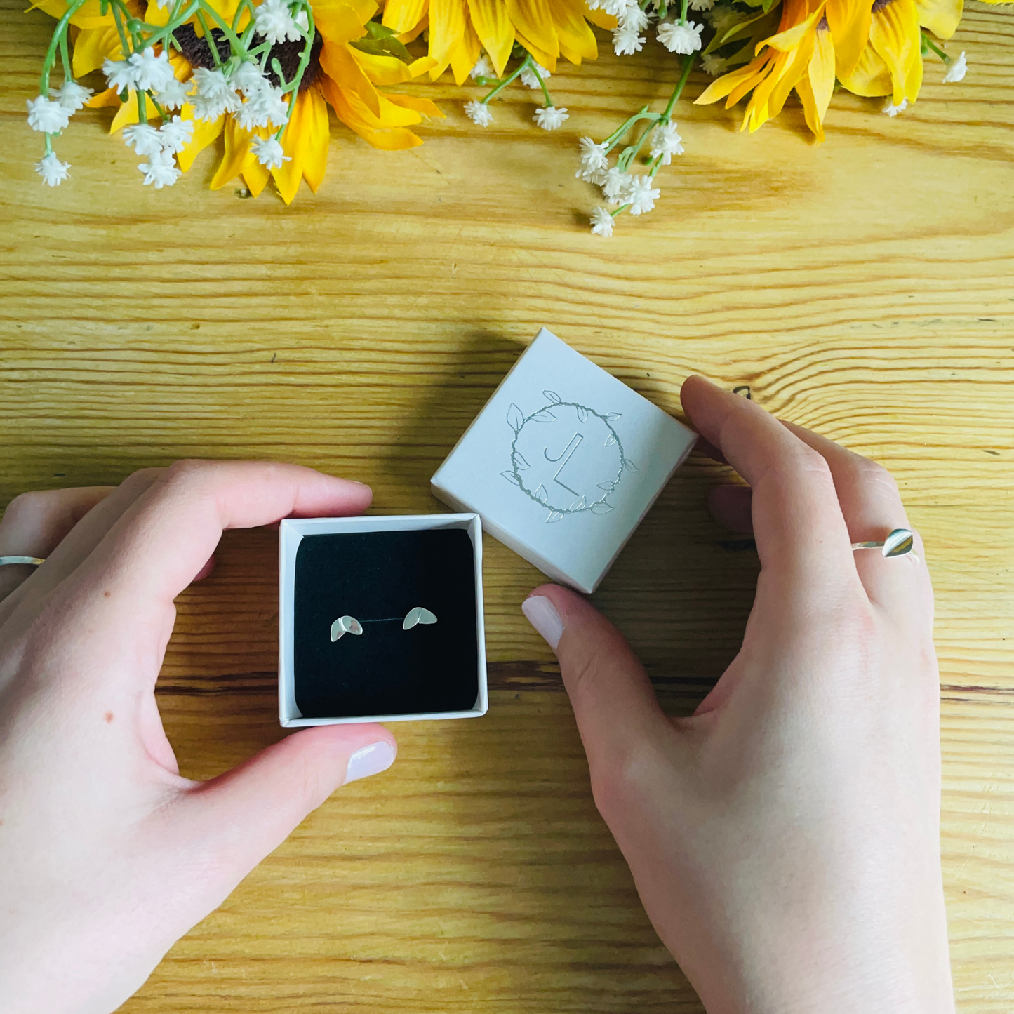 A pair of silver two leaf studs displayed in an ivory box with black insert. The box is being held in a pair of hands. One leaf on each stud is printed with a leaf print and the other is a shiny finish. Handmade by Jen Lithgo Jewellery