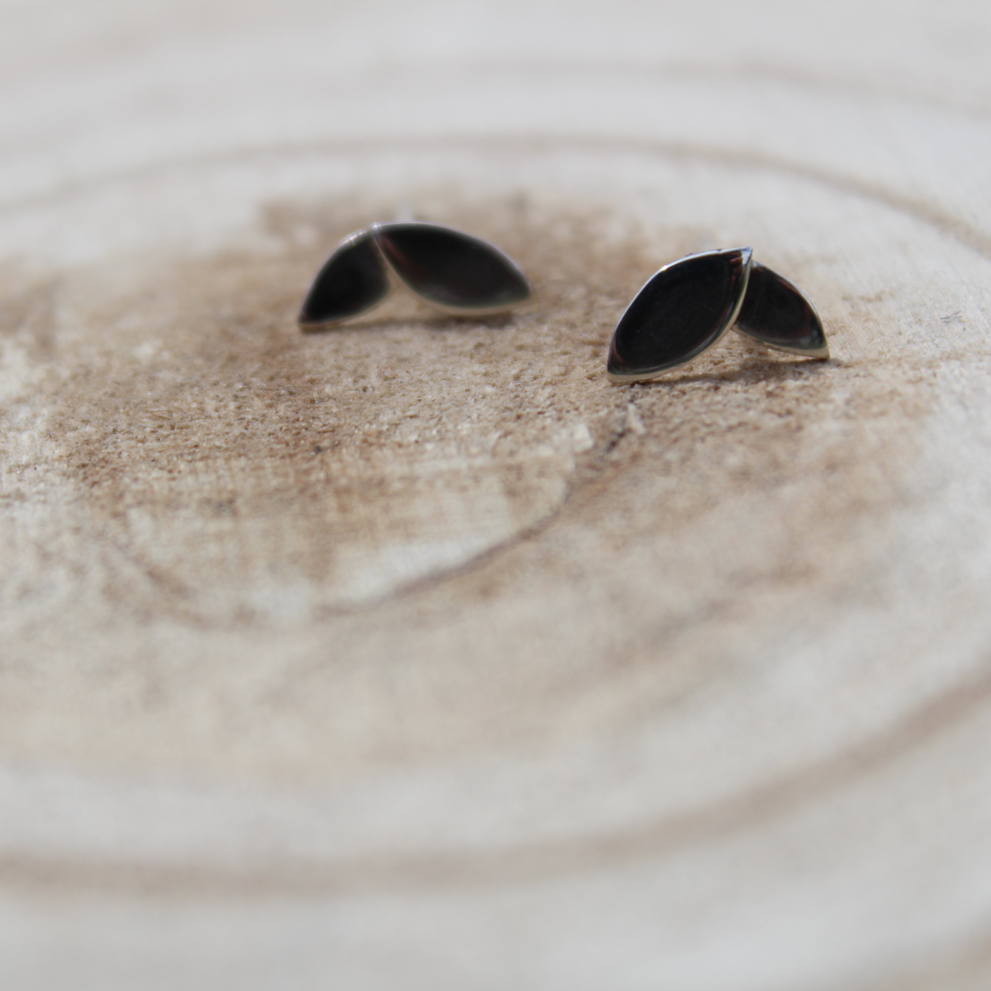 A pair of shiny finished two leaf stud earrings sat on a wooden plate. Handmade in sterling silver by Jen Lithgo Jewellery