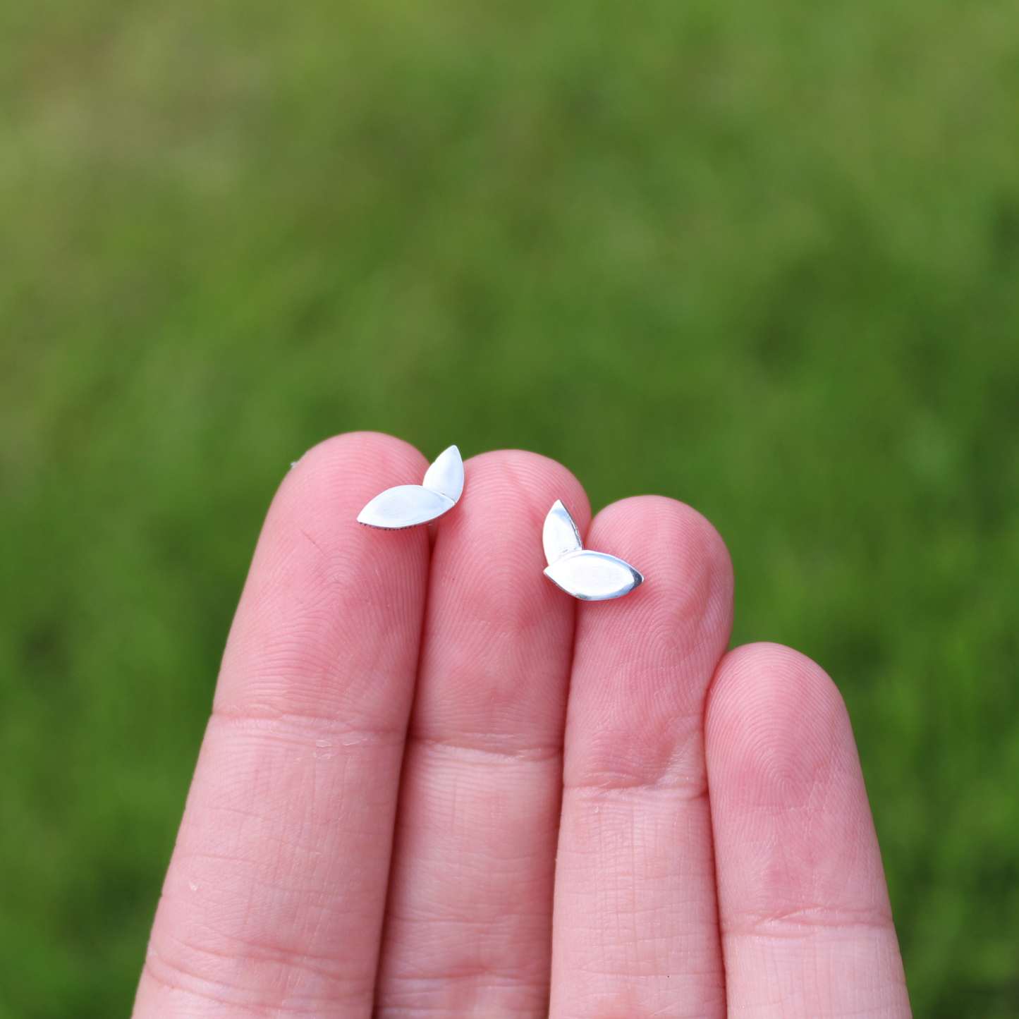 A pair of sterling silver two leaf stud earrings with a shiny finish. The studs are sat in between fingers, against a grassy background. Handmade by Jen Lithgo Jewellery