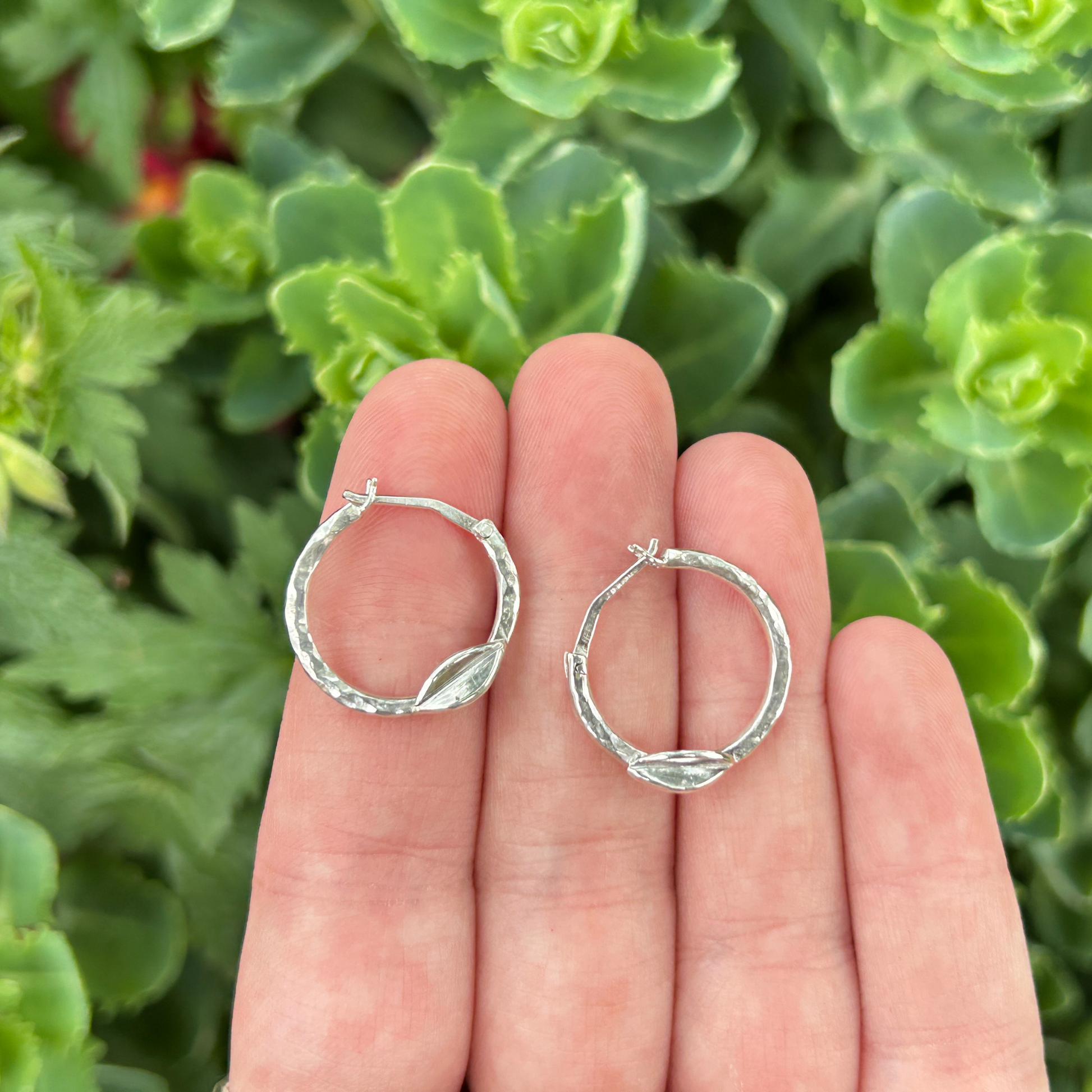 Handmade hammered sterling silver hoop earrings with leaf detail held against a background of green succulent plants – artisan nature-inspired jewellery