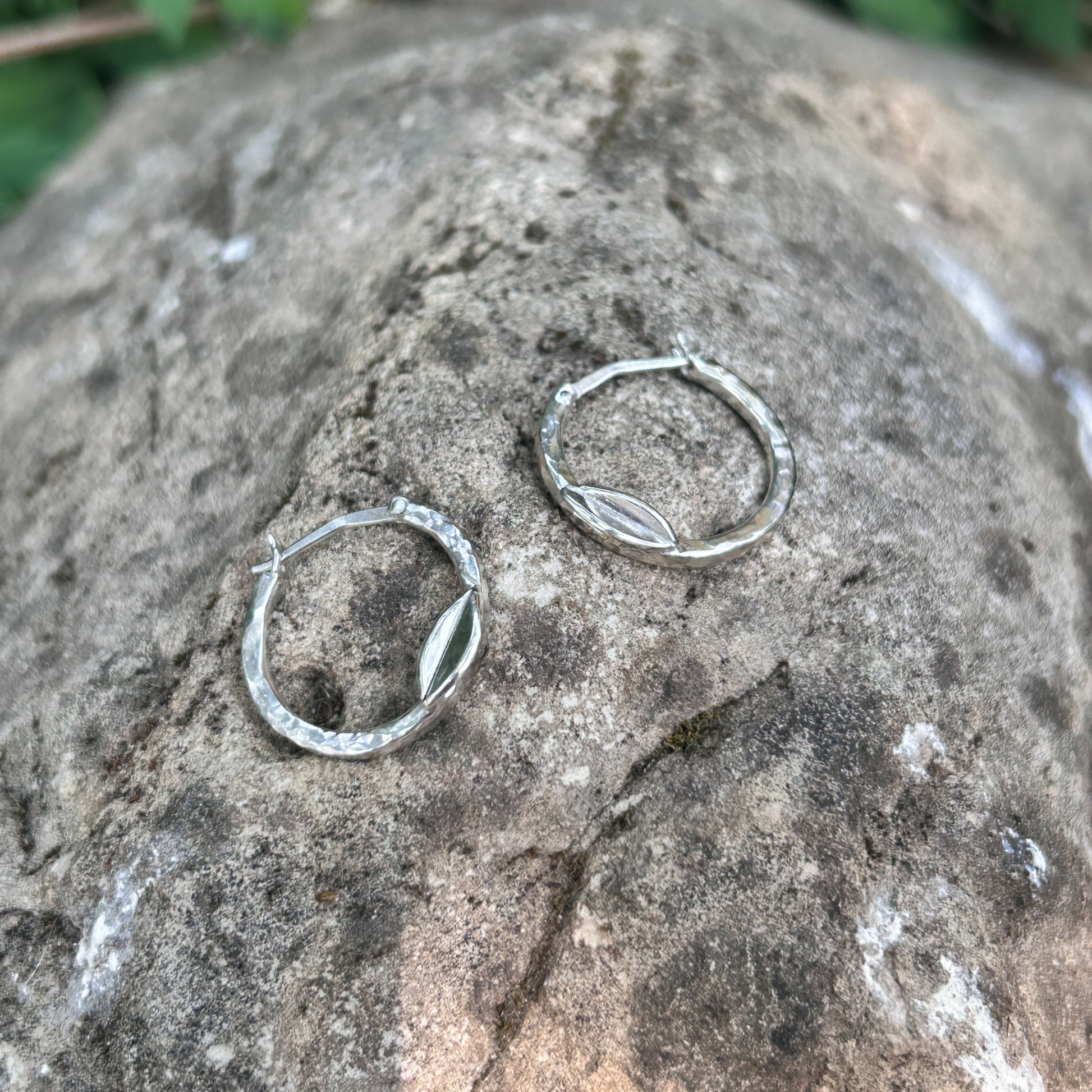 Handcrafted sterling silver hoop earrings with hammered texture and delicate leaf accent, displayed on natural stone background – eco-friendly artisan jewellery.