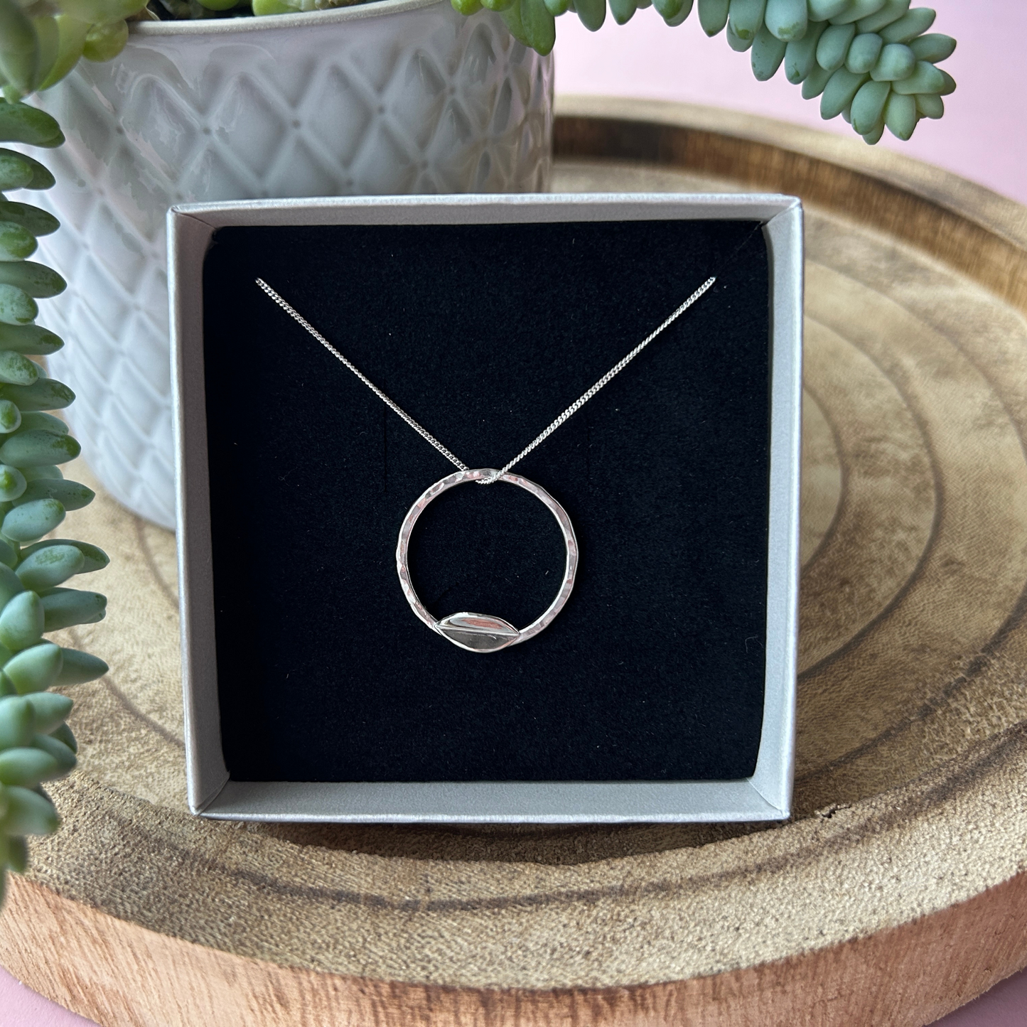 A silver necklace with a minimalist circular pendant is displayed in a black velvet-lined gift box. The pendant features a small leaf shaped accent at the bottom of the circle. The box rests on a wooden tray with a textured ceramic pot containing a green succulent in the background. The setting has a soft pink surface beneath the tray.