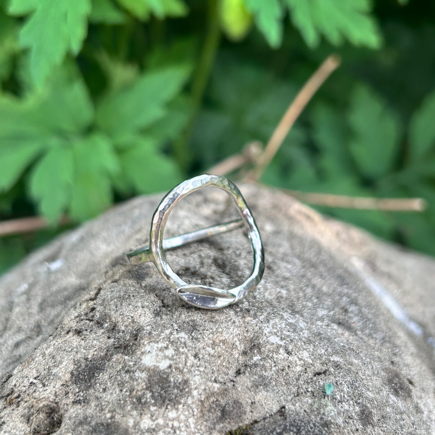 Handcrafted sterling silver circle ring with hammered texture and minimalist leaf detail, displayed on a natural stone background with green foliage. Eco-inspired artisan jewellery for nature lovers.