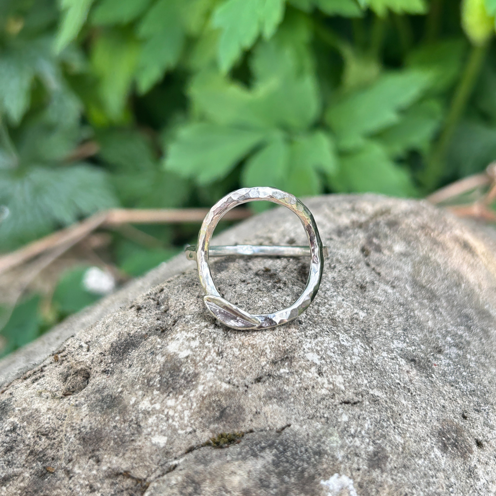 Handcrafted sterling silver circle ring with hammered texture and minimalist leaf detail, displayed on a natural stone background with green foliage. Eco-inspired artisan jewellery for nature lovers.