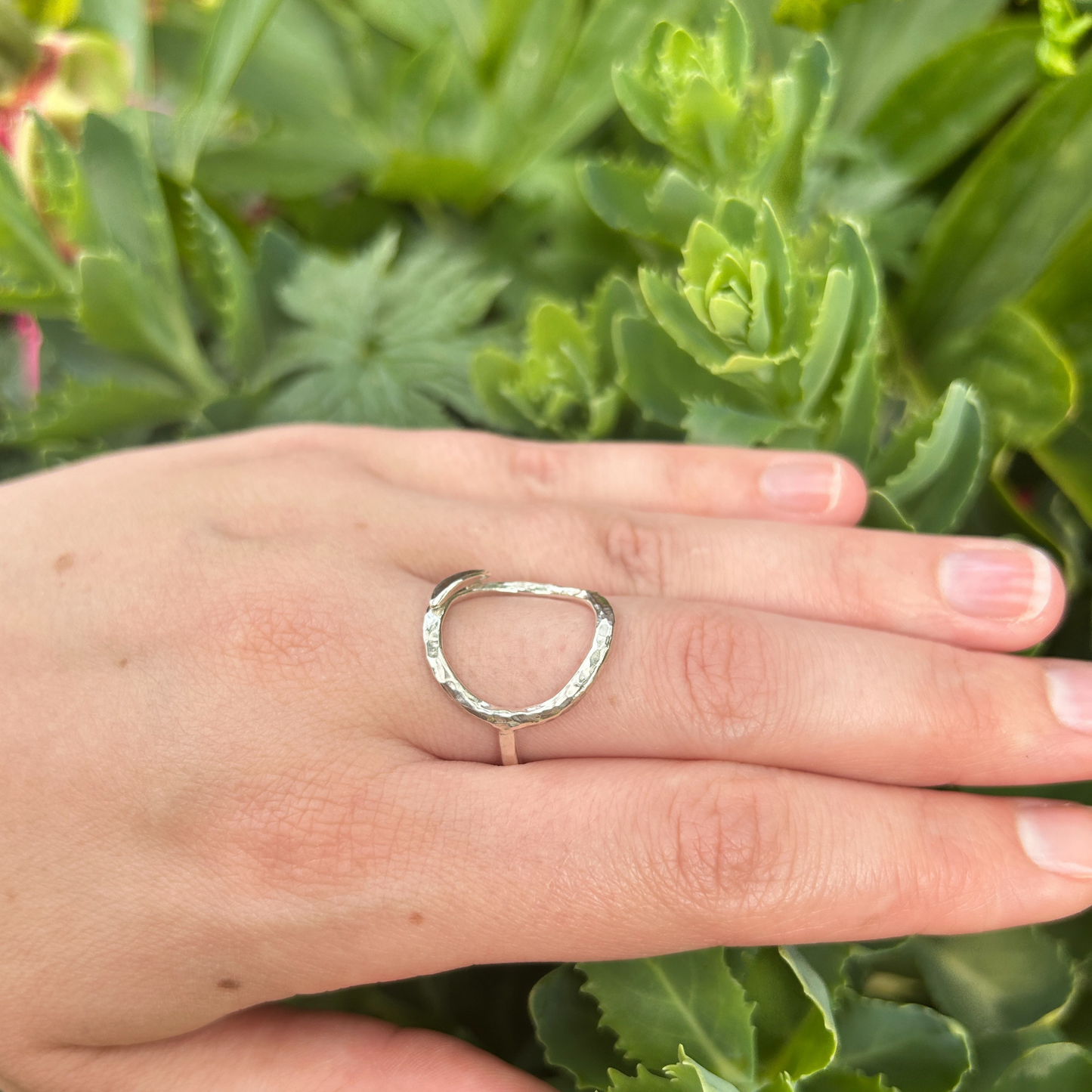 A side view of a hand wearing a hammered open circle silver ring with a succulent in the background