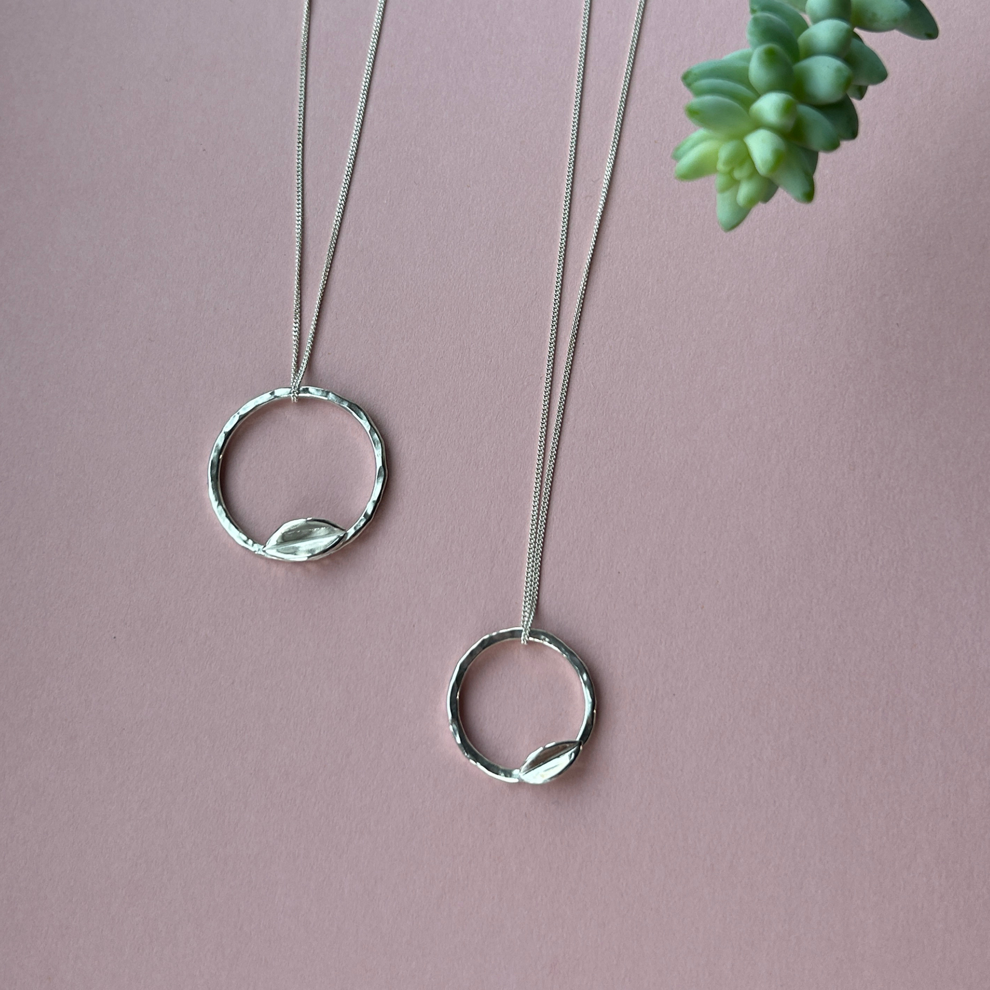 Two sizes of Hammered effect hoop leaf necklaces laid side by side on a pink background with a small piece of donkey tail succulent in the corner of the image. Handmade in sterling silver by Jen Lithgo Jewellery
