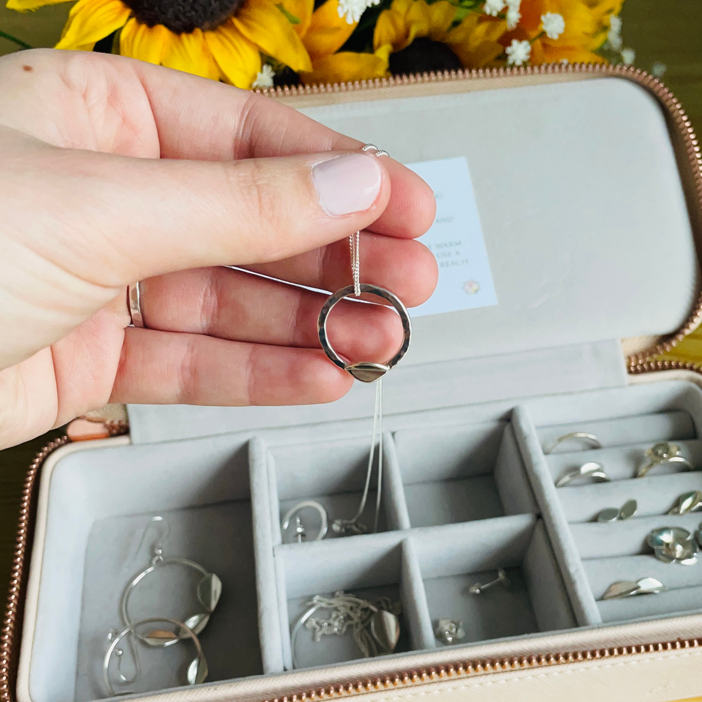 A small hammered effect hoop leaf necklace hanging on an 18" sterling silver chain in a hand over a jewellery box full of silver jewellery handmade by Jen Lithgo Jewellery