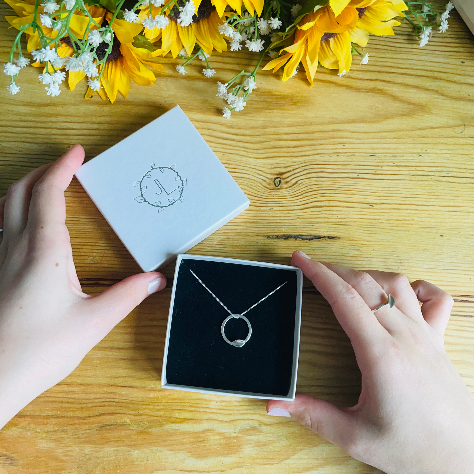 A silver hammered effect leaf necklace in an ivory branded box with black insert on a wooden table top - handmade by Jen Lithgo Jewellery