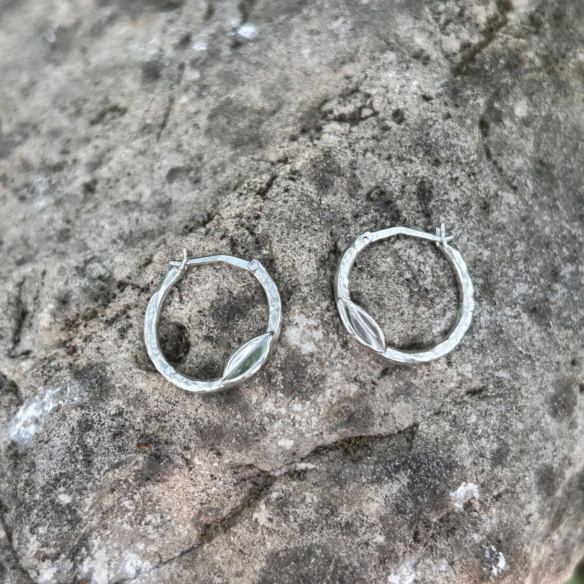 Handcrafted sterling silver hoop earrings with hammered texture and delicate leaf accent, displayed on natural stone background – eco-friendly artisan jewellery.