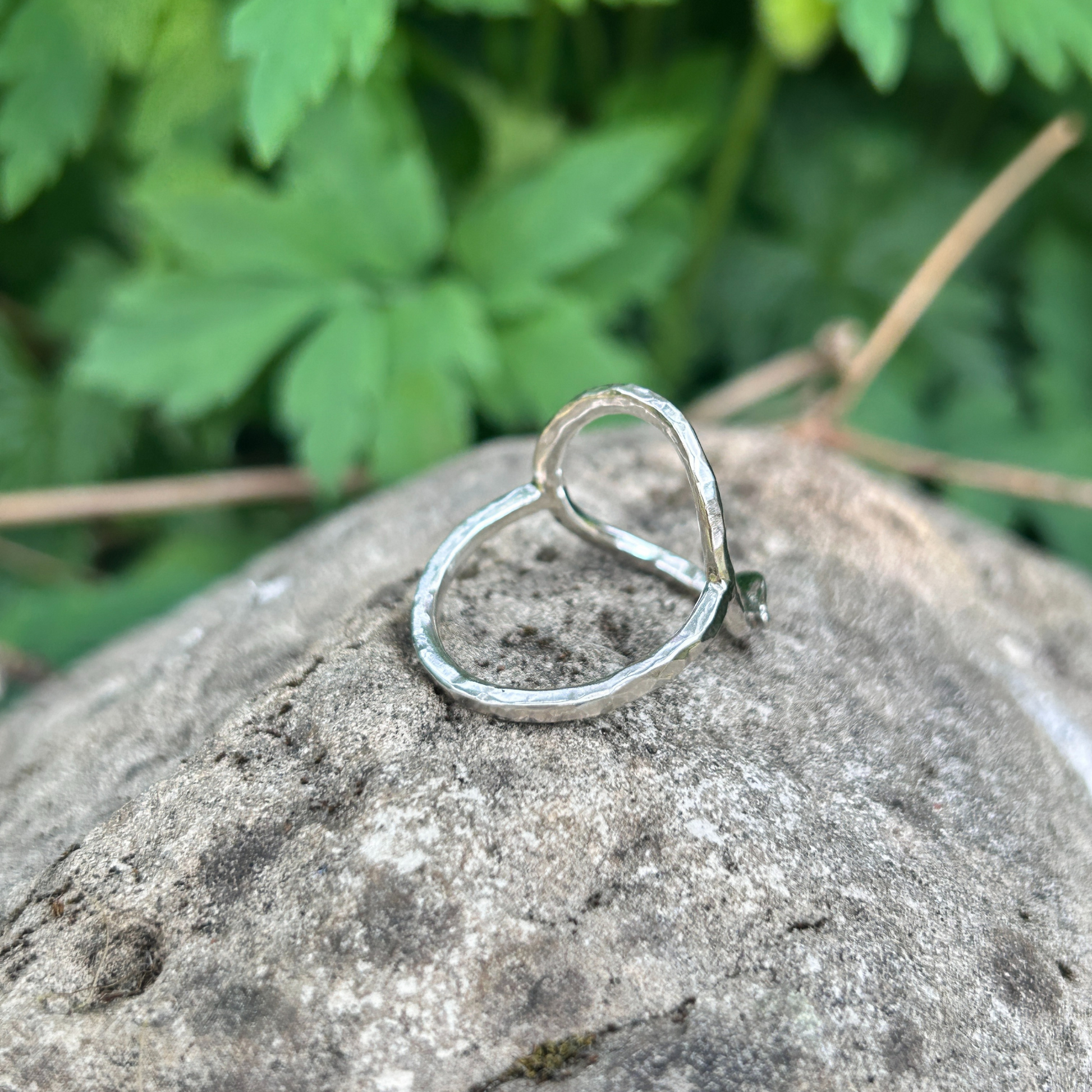 Side to rear view of a handcrafted sterling silver circle ring with hammered texture and minimalist leaf detail, displayed on a natural stone background with green foliage. Eco-inspired artisan jewellery for nature lovers.