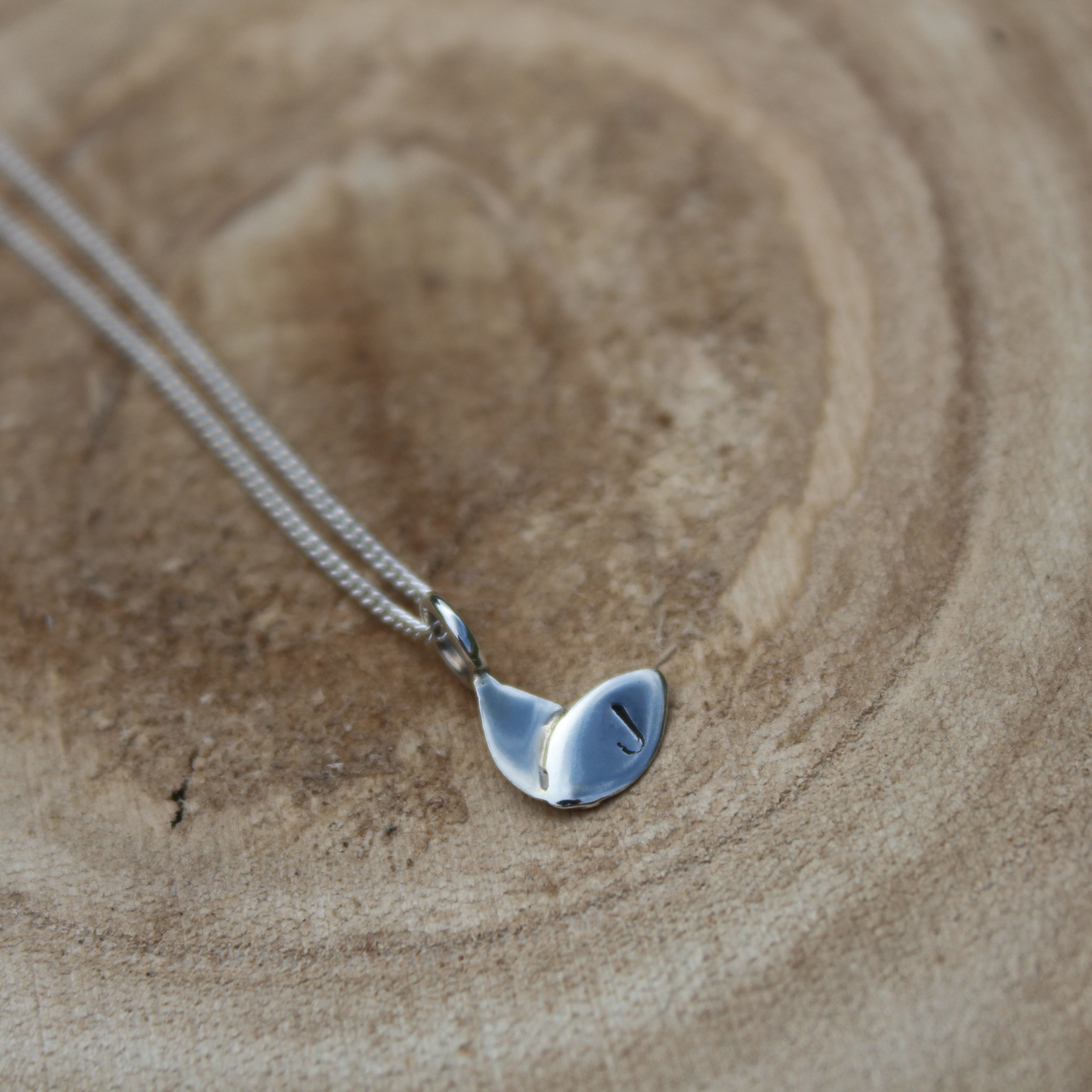 A silver duo leaf pendant personalised with a hand stamped 'J' on the right leaf laid on a wooden background. Handmade by Jen Lithgo Jewellery