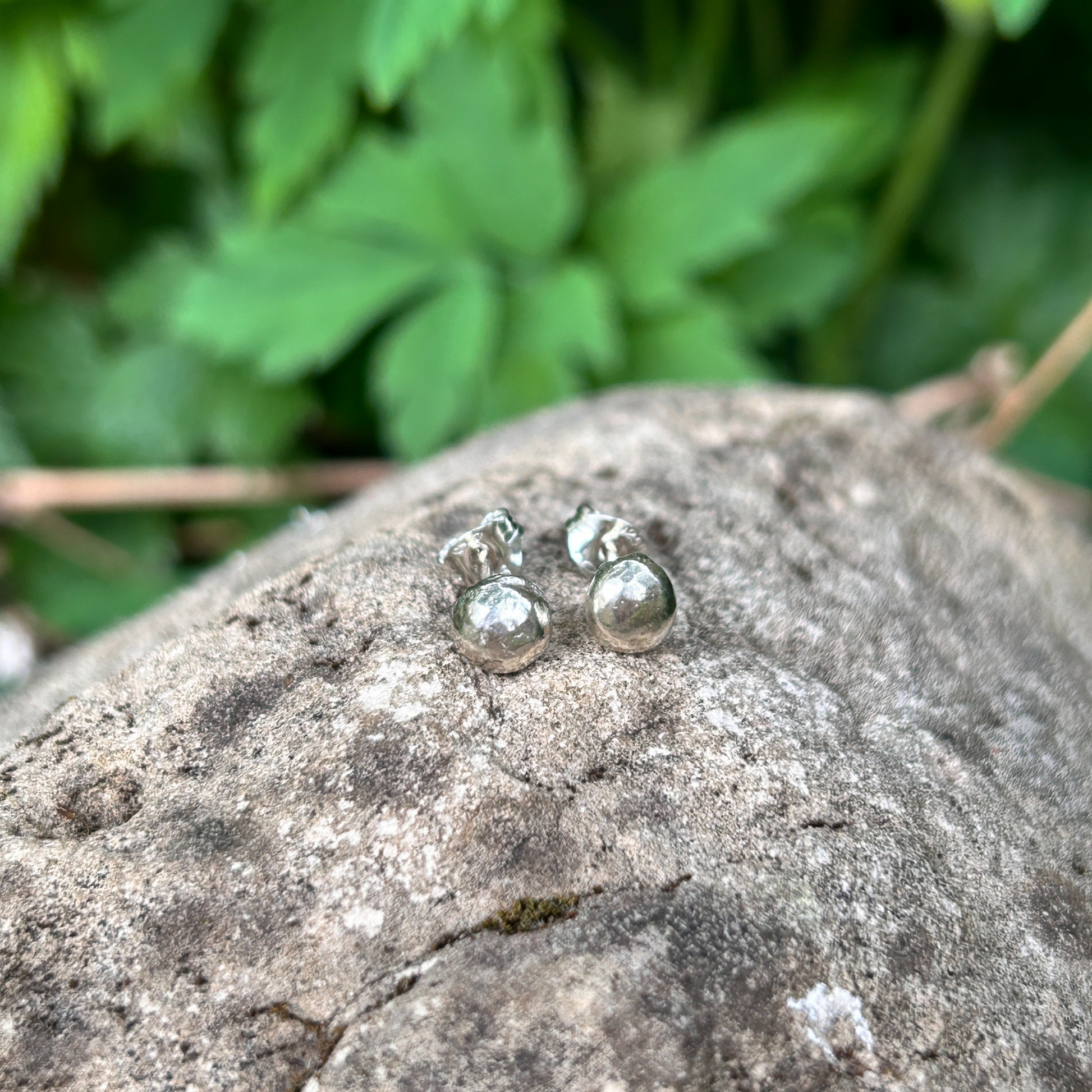 Large sterling silver pebble earrings sat on a rock with green foliage behind - handmade nature lover jewellery
