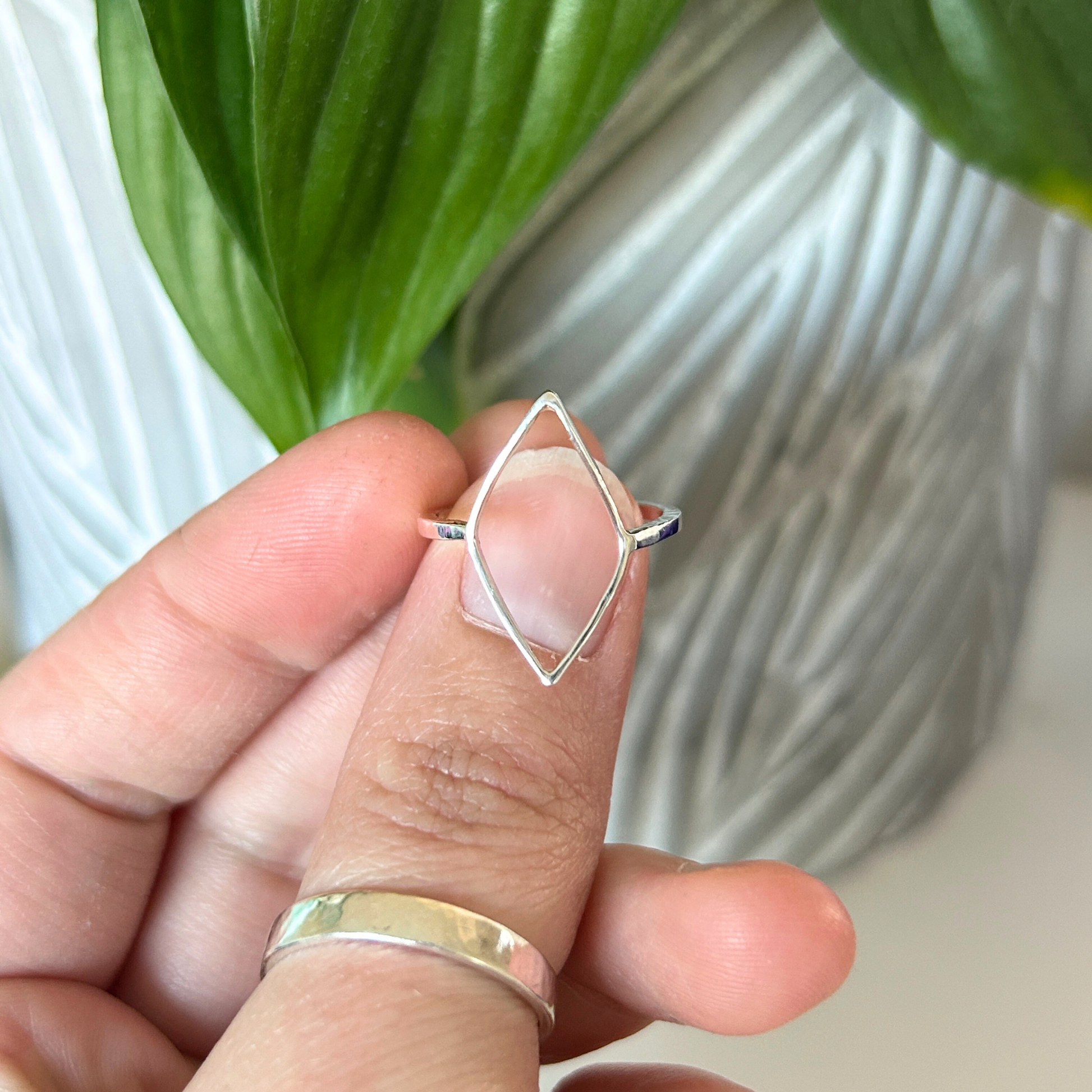 Hand holding a handmade sterling silver ring with a geometric open leaf design, blurred green plant in the background