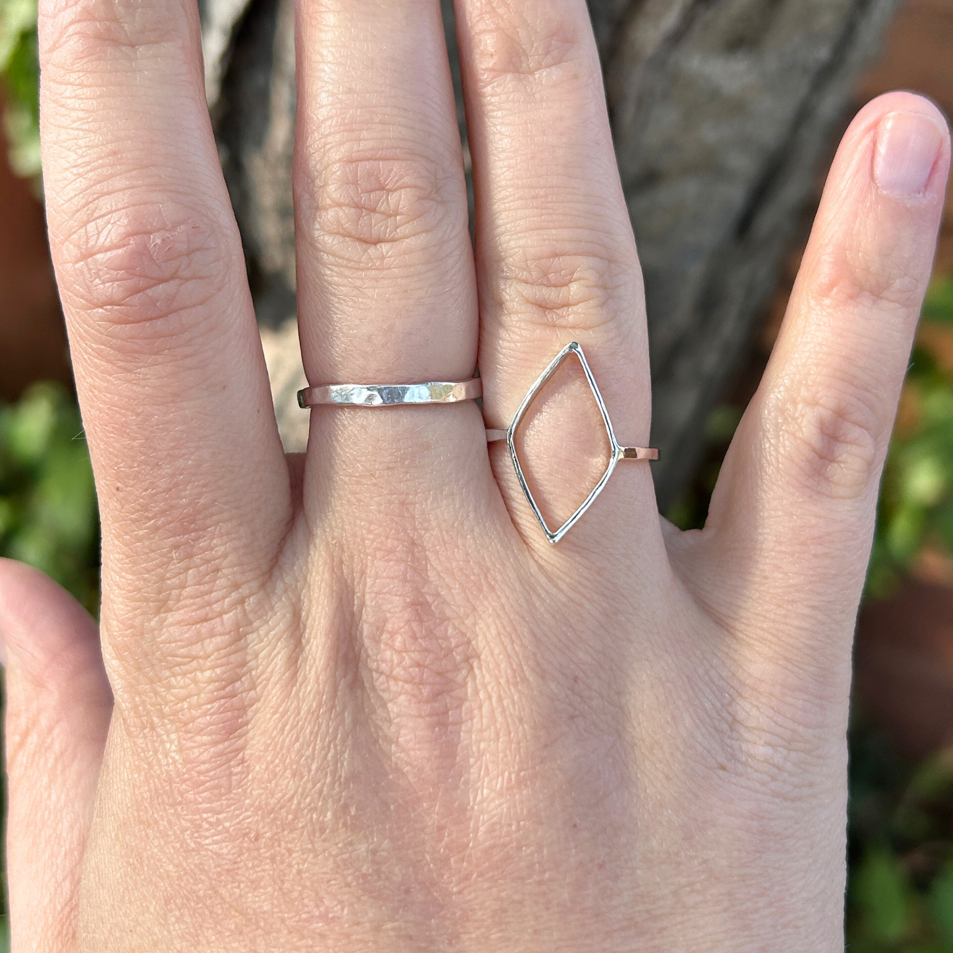 Hand wearing two rings - a hammered textured ring and a geometric open leaf ring, both handmade from 100% recycled sterling silver with a blurred natural background