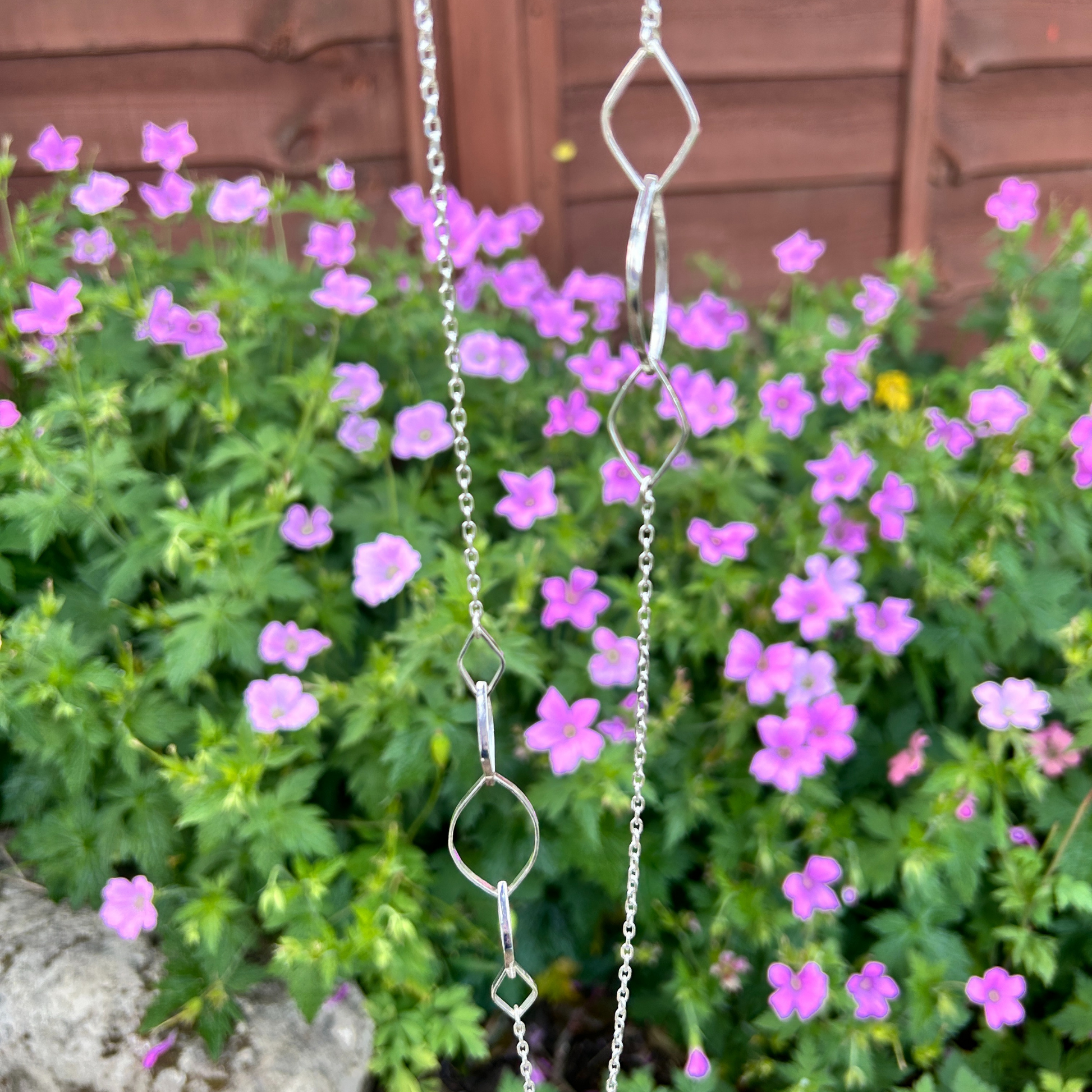 Refined sterling silver asymmetrical designer necklace featuring interlocking leaf shaped links on a delicate chain, hanging in front of pink flowers in a garden.