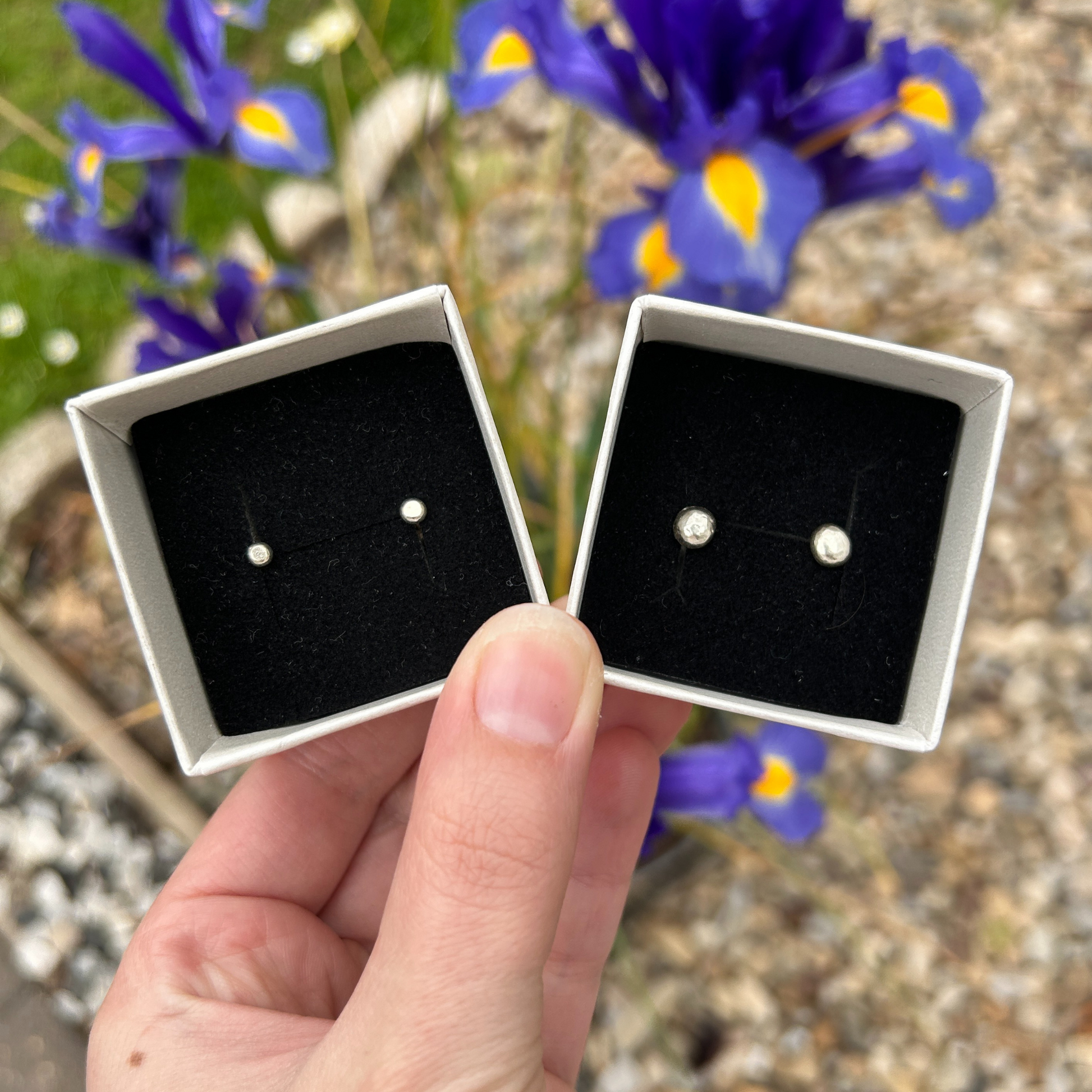 Handmade pebble stud earrings (small and large) in a box with black velvet being held over some purple irises.
