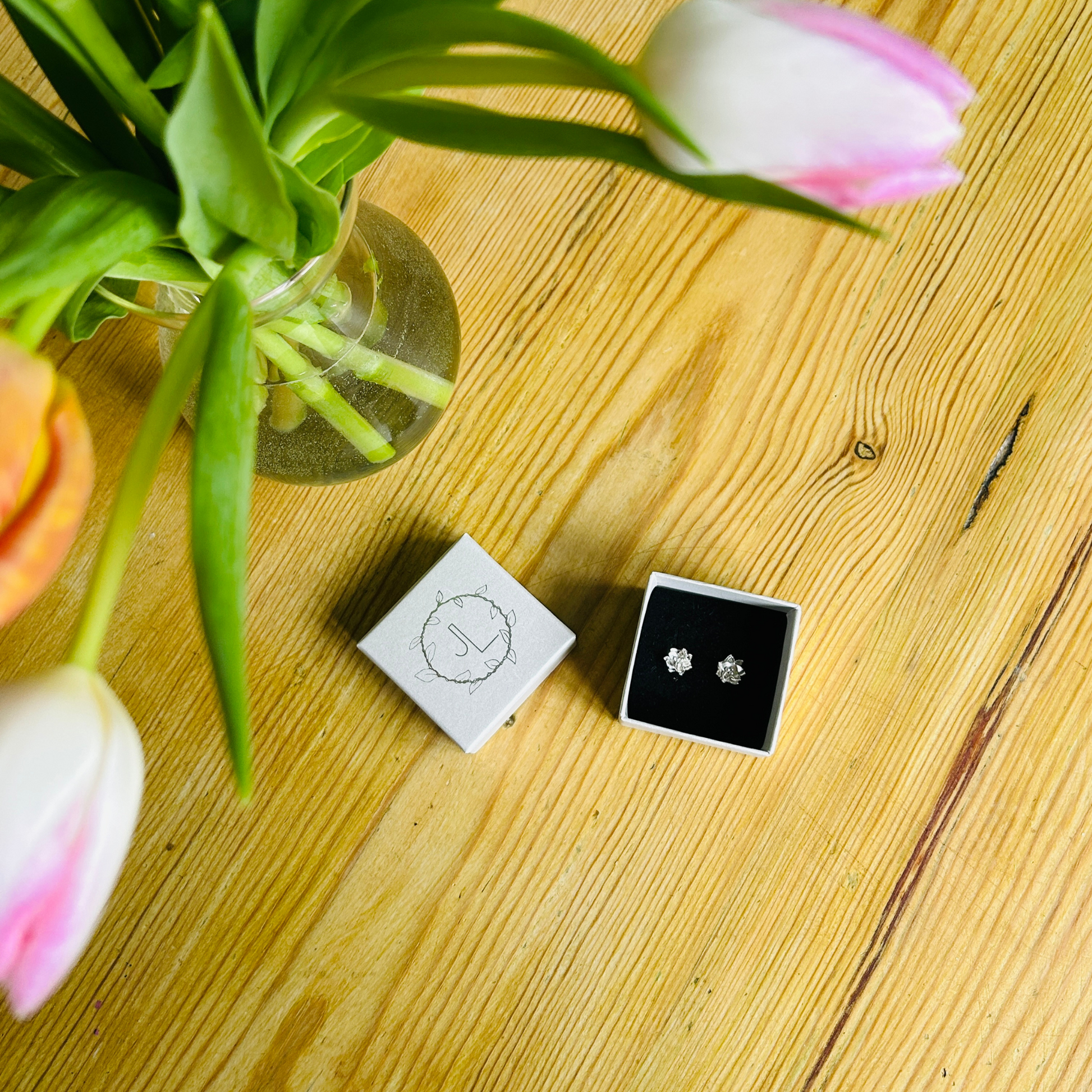 An open box displaying a pair of sterling silver flower stud earrings on a black velvet insert. Off to the top left corner, there are some pink and white tulips in a clear vase. Background is a wooden table top. Handmade stud earrings by Jen Lithgo Jewellery