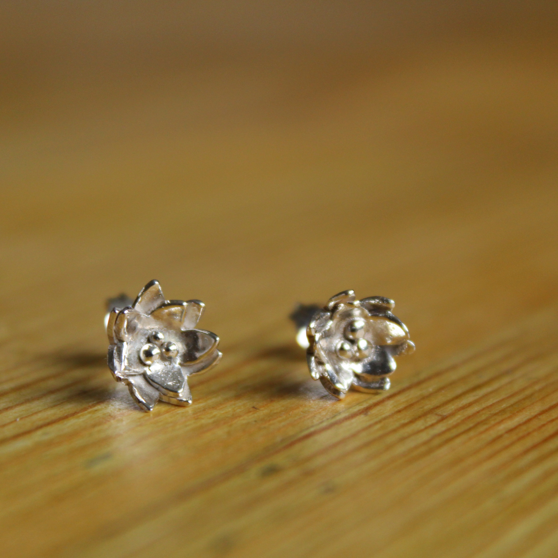 A pair of sterling silver flower earrings in the shape of water lilies sitting on a wooden table top. Handmade by Jen Lithgo Jewellery