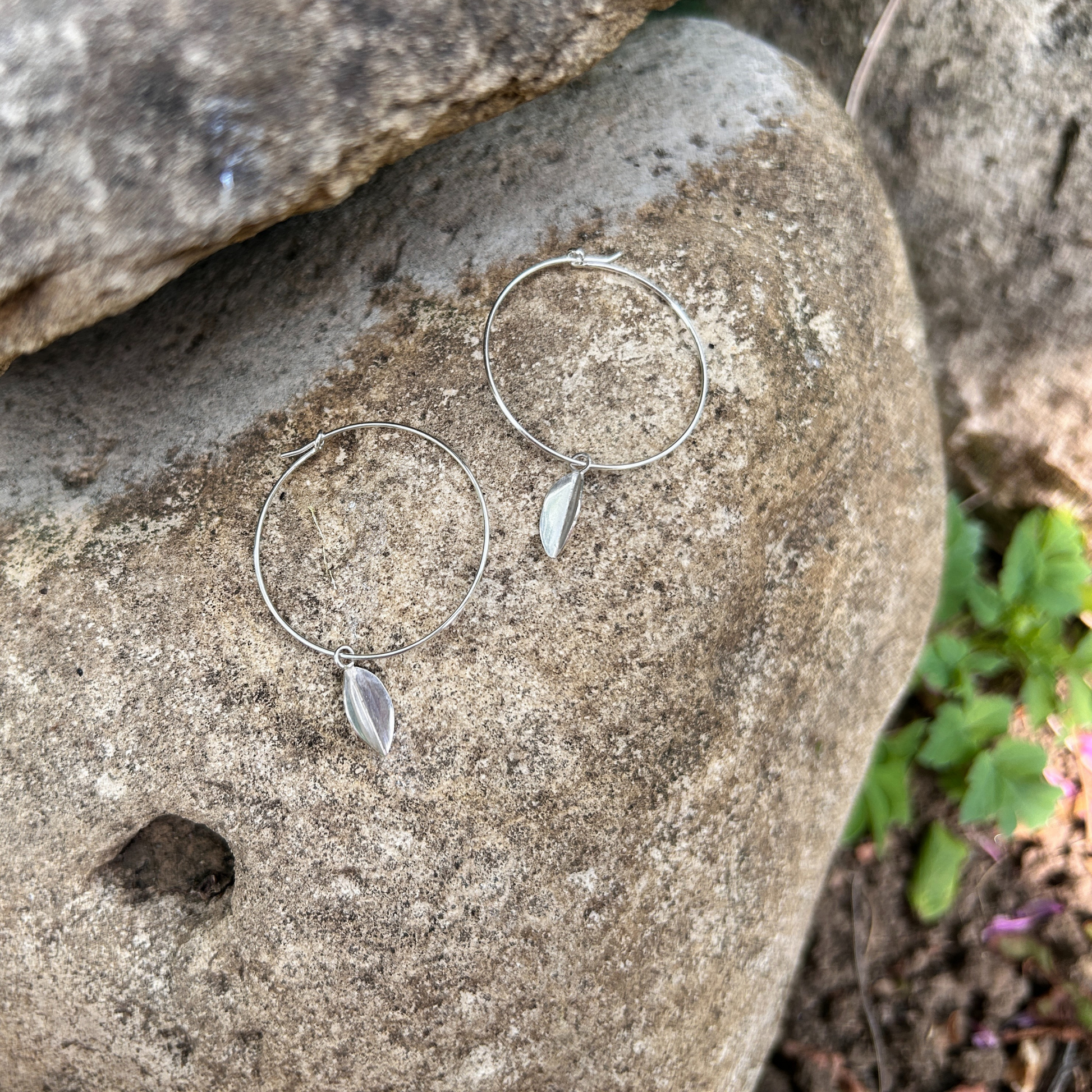 Wire hoop earrings laid on a stone background with a silver leaf hanging from each hoop.