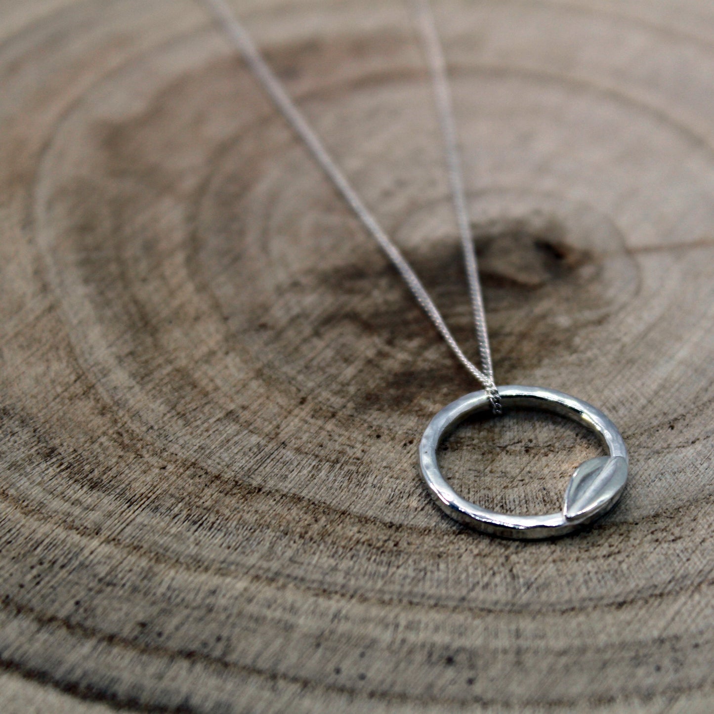 A silver necklace with a minimalist circular pendant is displayed on a wooden background. The pendant features a small leaf shaped accent at the bottom of the circle and the pendant hangs from an 18" sterling silver chain. Handmade by Jen Lithgo Jewellery