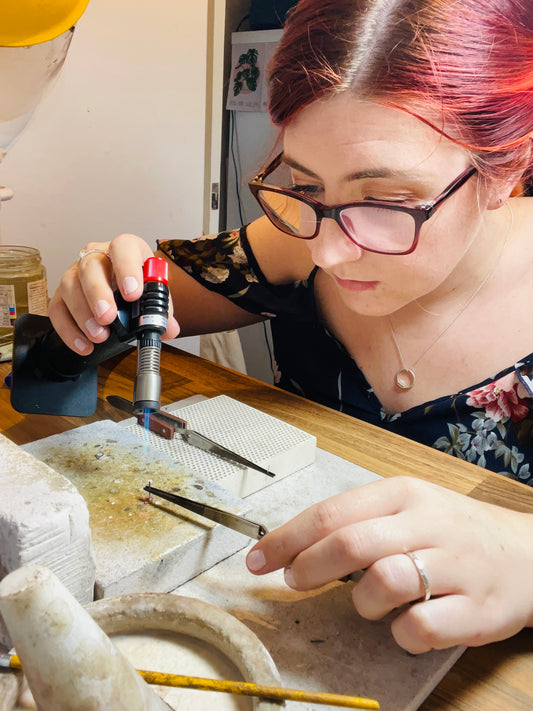 Jen Lithgo holding a small blowtorch and tweezers over a soldering block. Soldering a post onto an earring. Jen has red hair, wears red and black glasses and a blue floral cami top.