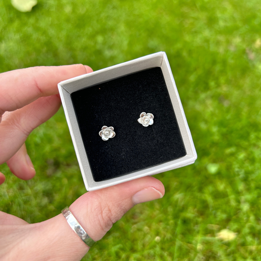A hand holding dainty flower stud earrings in a pearlescent ivory box with black insert with a grass