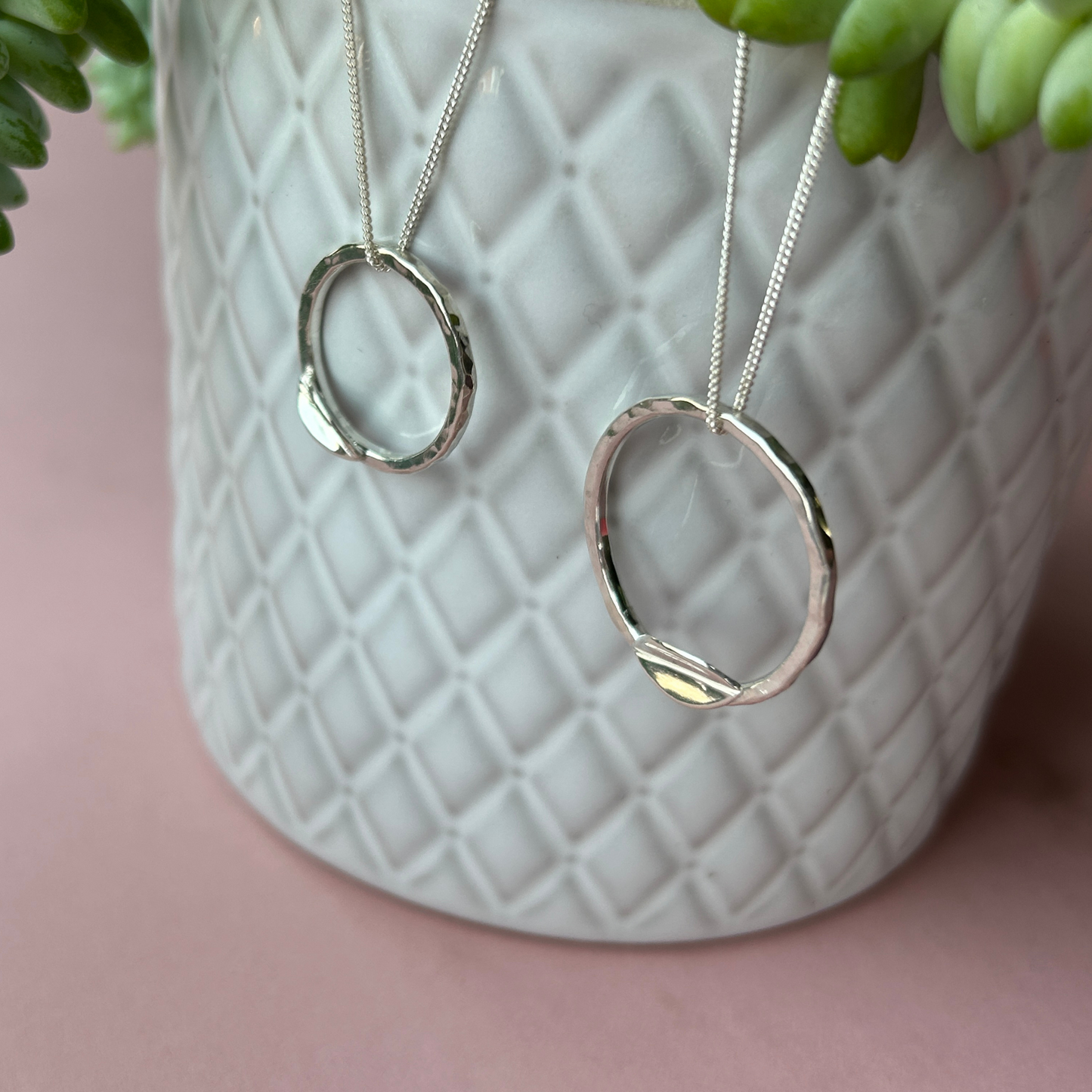 Two handmade sterling silver hammered hoop leaf necklaces hanging on a grey textured plant pot. Jen Lithgo Jewellery 