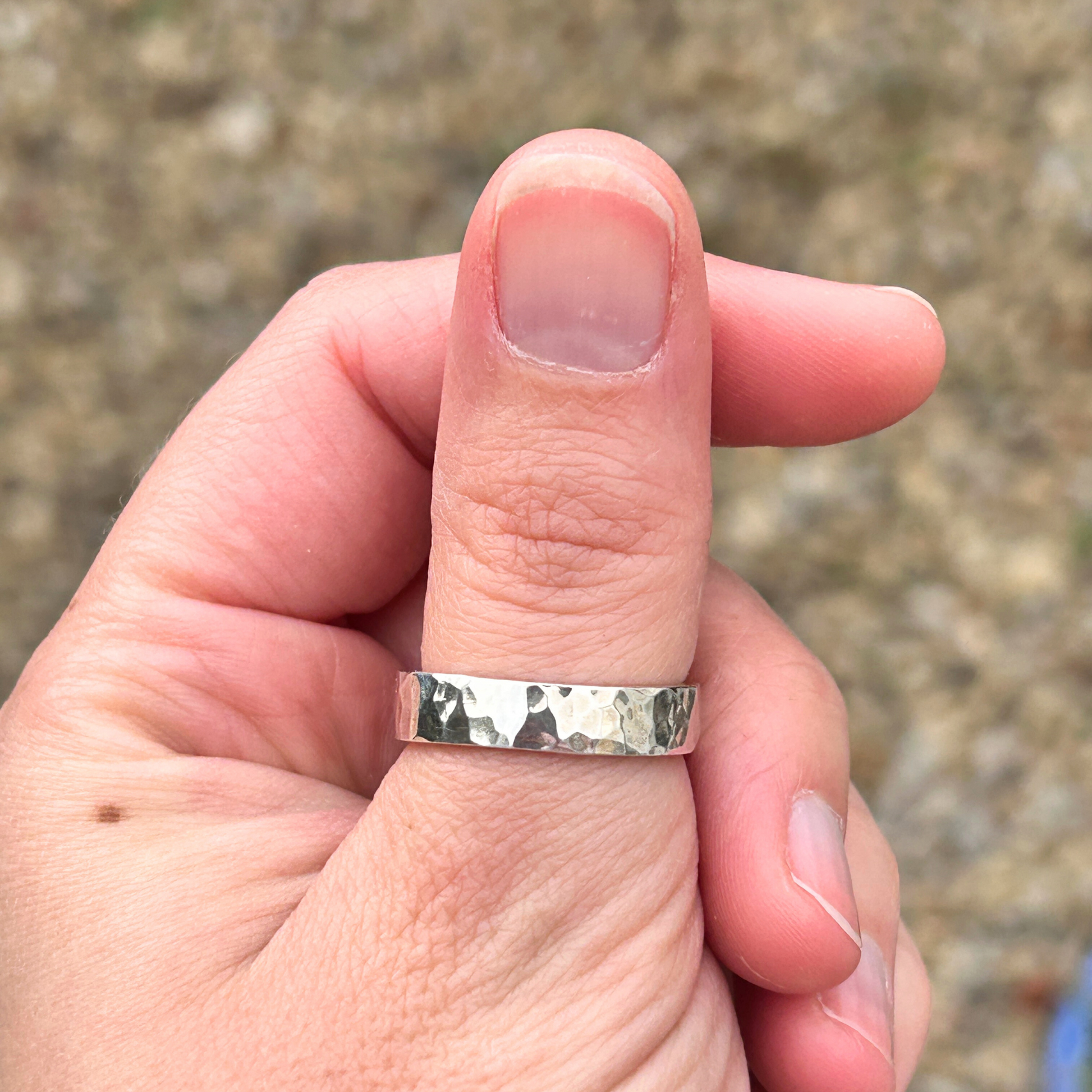 Hand wearing a hammered 5mm sterling silver ring on a thumb with a textured band against a blurred natural background