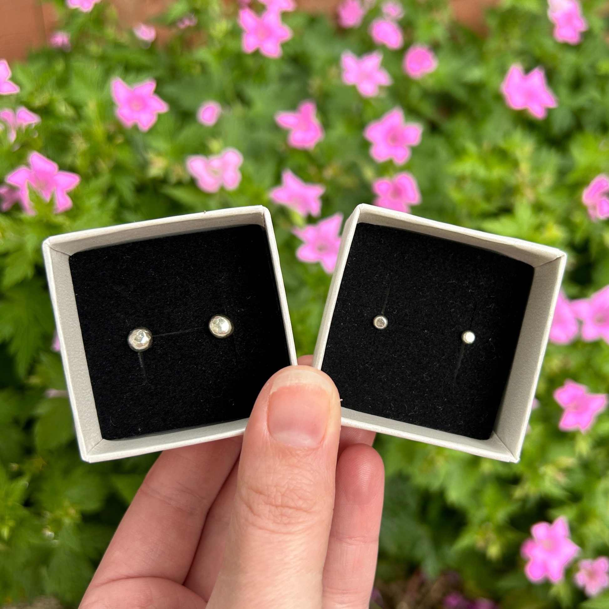 Large & small pebble stud earrings in a box on black velvet being held over a flower bed 