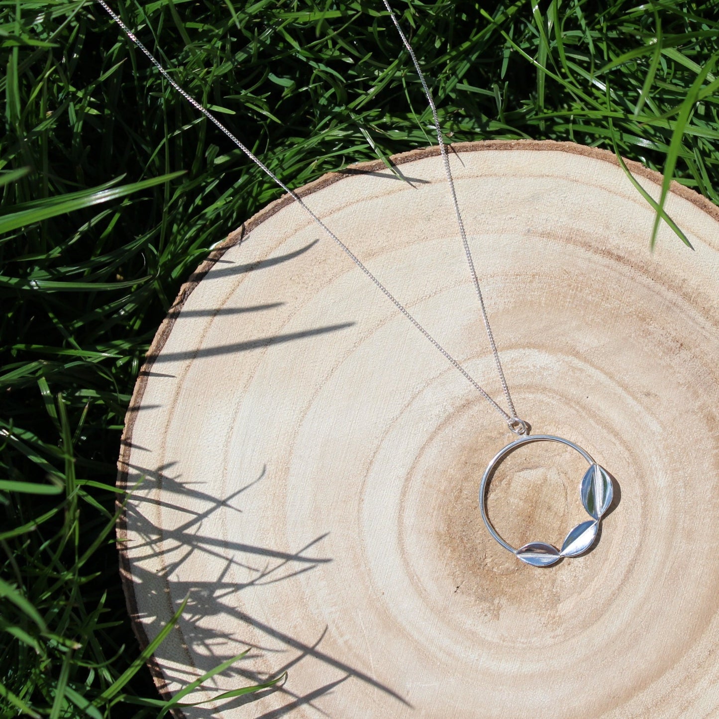 Wooden log on grass with sterling silver three leaves on a hoop necklace hanging on a curb chain, handmade by Jen Lithgo Jewellery