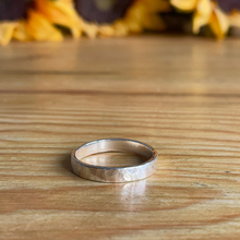Load image into Gallery viewer, Close up of Hammered Effect 4mm Ring, handmade in sterling silver and sat on a wooden table top with sunflowers in the background. Handmade by Jen Lithgo