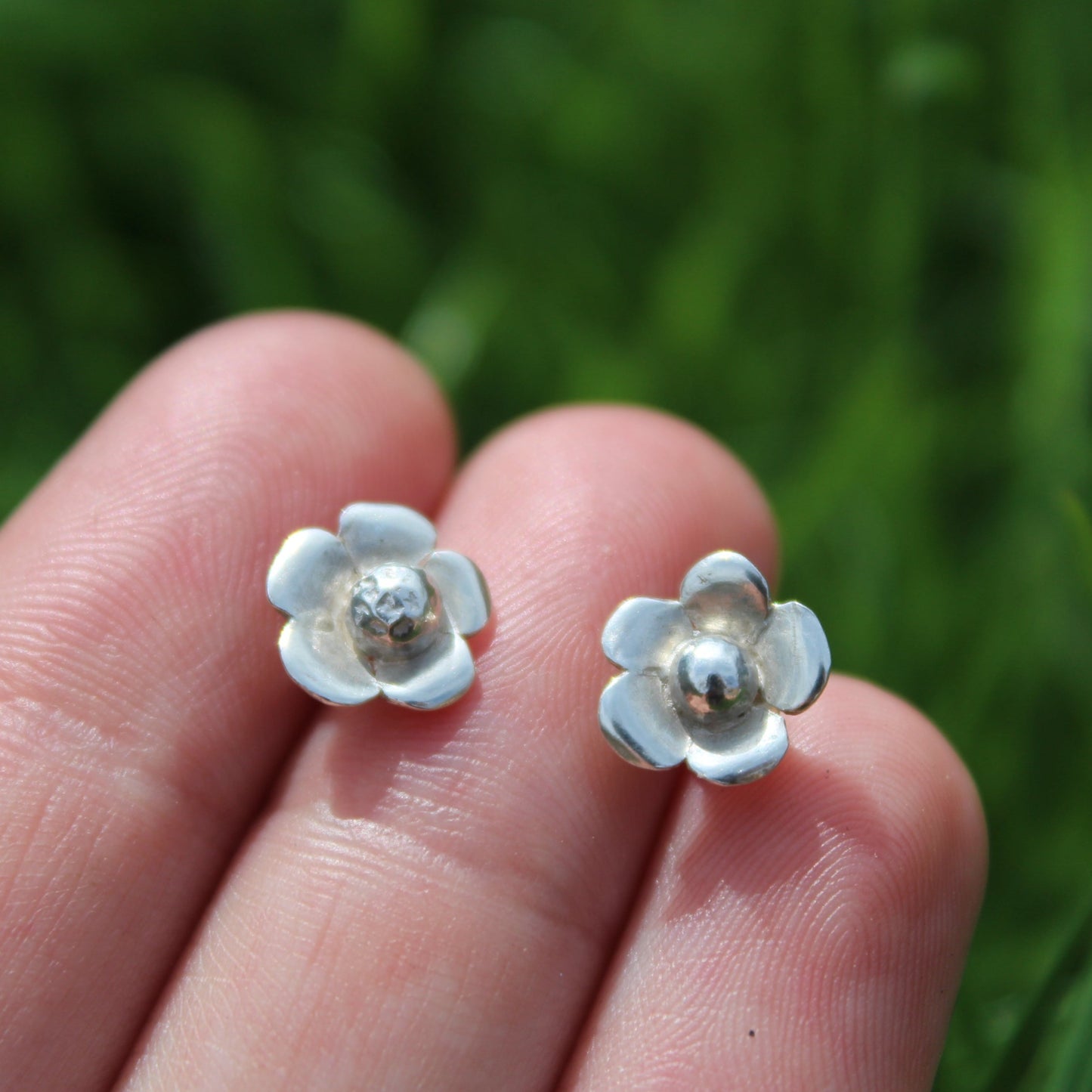 A pair of silver flower stud earrings held in fingers with a grassy background