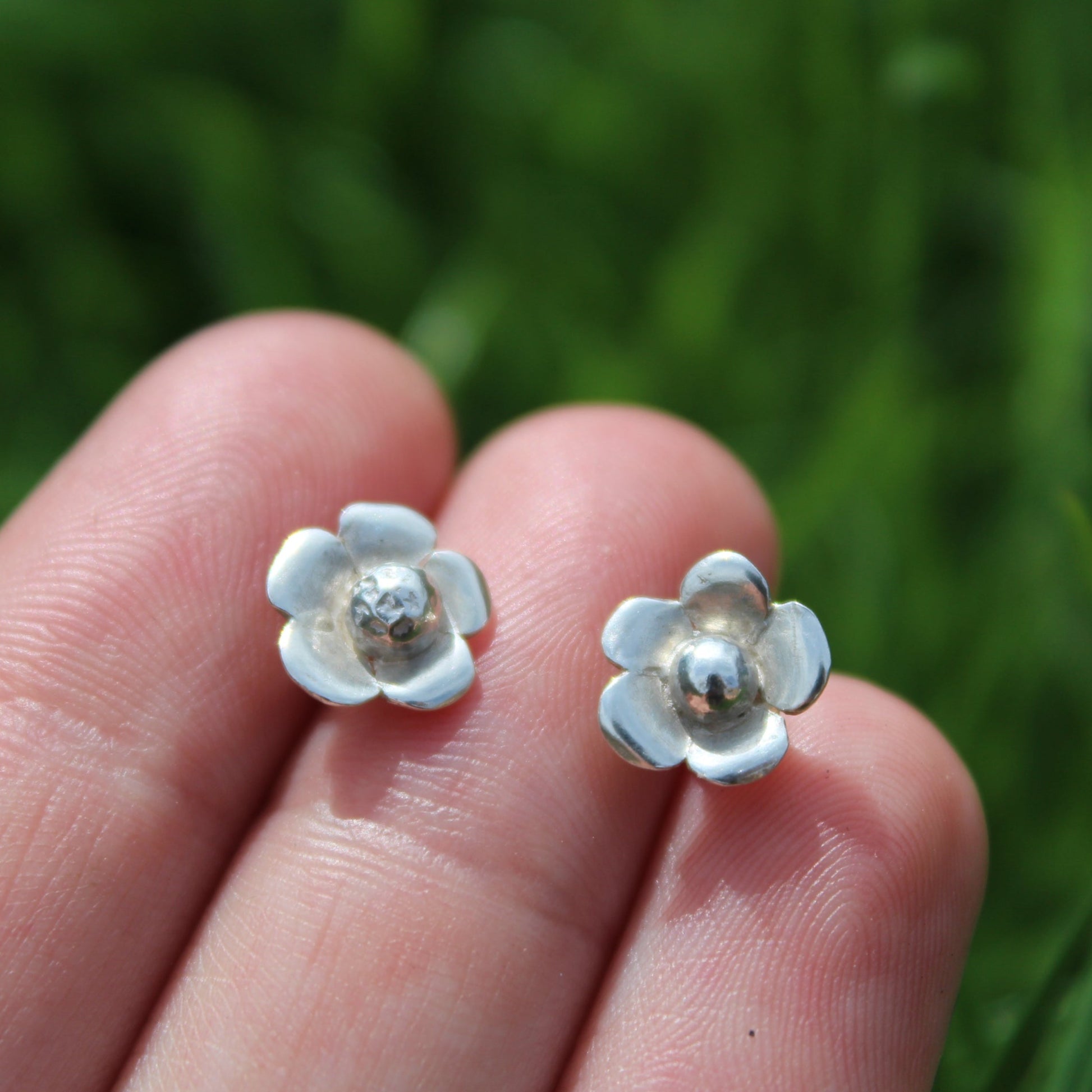 A pair of silver flower stud earrings held in fingers with a grassy background