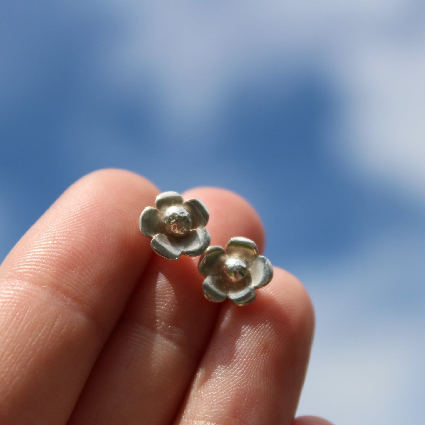 A pair of silver flower stud earrings held in between fingers against a sky background